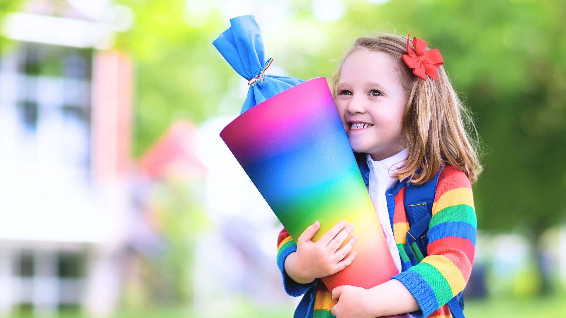 Kind Mädchen mit Regenbogenzuckertüte in der Hand steht vor einer Schule Einschulung
