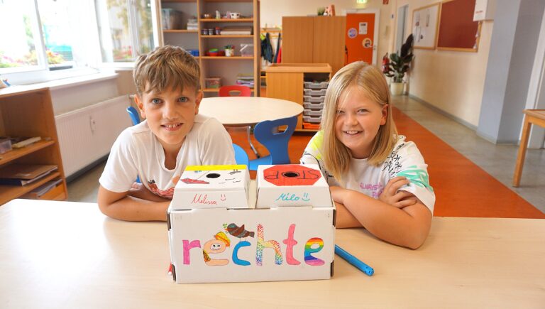 Weltkindertag in Radebeul zwei Kinder sitzen vor einer Papkiste auf dem das Wort "Rechte" steht. Die Kiste hat zwei Löcher und Melissa und Milo steht drauf