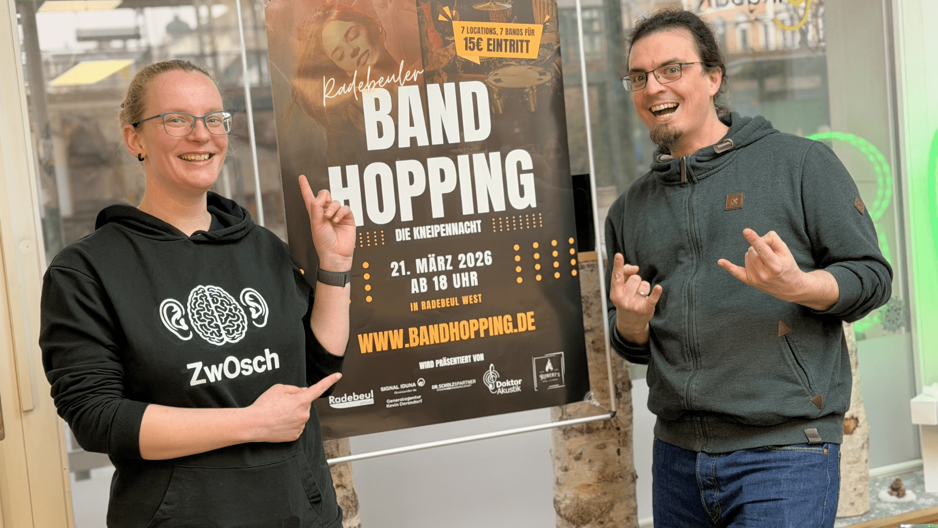 Daniel und Theresa Doktor stehen vor dem Band Hopping Plakat in ihrem Hörakustikladen in Radebeul West